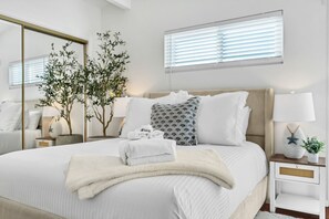 Ein ordentlich eingerichtetes Schlafzimmer mit einem großen Bett, einem Nachttisch mit Lampe, einem Spiegel und einem Fenster mit Jalousien.
