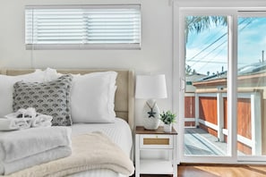 A bedroom with a bed, a nightstand, a lamp, and a view of a wooden deck through a sliding glass door. - Ocean Beach Chic 2BR | Patio | Surf Vibes | A/C (San Diego)
