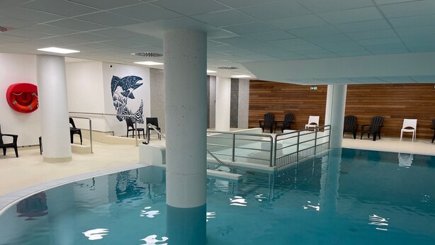 Piscina interior com superfície de água transparente, rodeada por um deck com guarda-corpo de metal. Há cadeiras e um salva-vidas no deck.