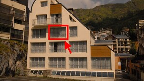Um edifício com várias andares e varandas, uma área de estacionamento na frente e montanhas ao fundo.