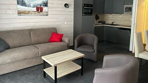Una sala moderna con un sofá, una mesita y una silla. Se ve una cocina al fondo con armarios oscuros y electrodomésticos modernos.