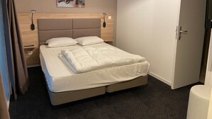 Una cama bien hecha con cabecera, una mesita de noche con lámpara, una obra de arte en la pared y una puerta a la derecha.