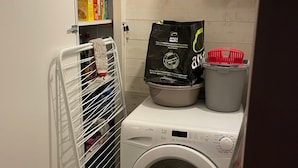 Kompakti pyykinpesutila, jossa on etuaukollinen pesukone, pyykkiteline, kori ja pussi pyykinpesuainetta.