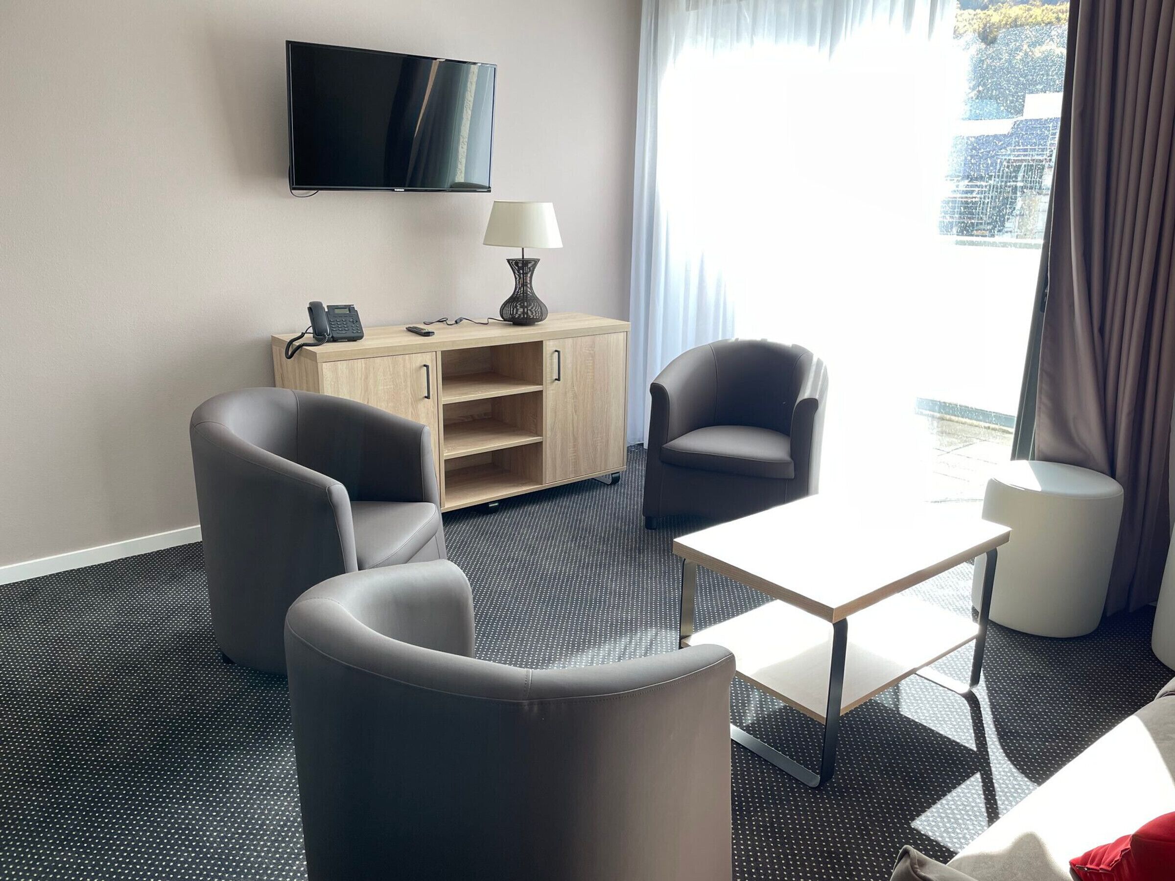 A hotel room with a flat-screen TV mounted on the wall, a wooden cabinet with a lamp, two grey armchairs, a small white table, and a view of the city through large windows.