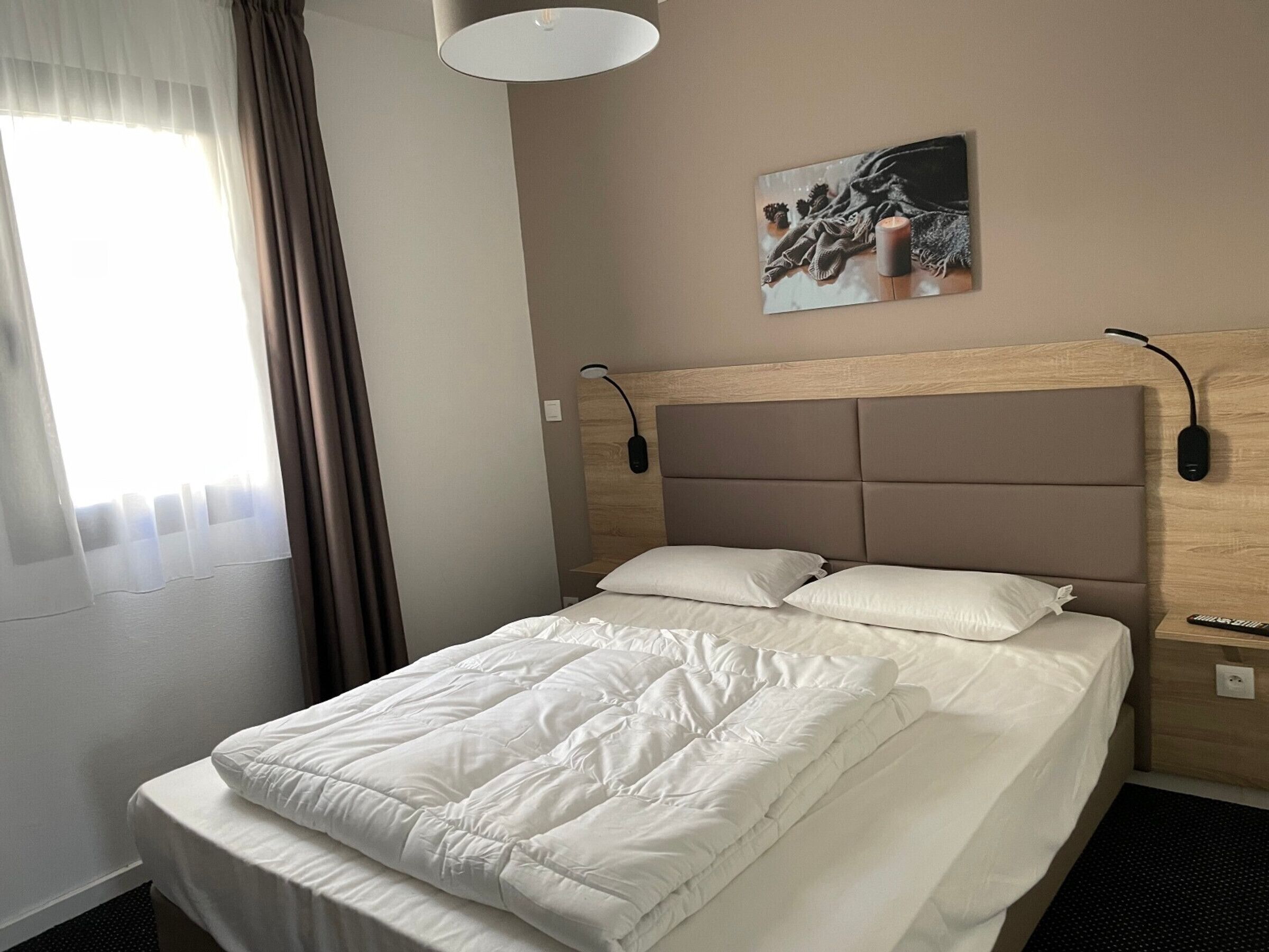 A hotel room with a bed, a nightstand, a lamp, a painting on the wall, and a window with curtains.