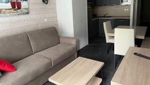 Ett modernt vardagsrum med en soffa, ett soffbord och en matplats med ett bord och stolar. I bakgrunden syns ett kök med en mikrovågsugn och skåp.