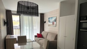 Ett kompakt hotellrum med ett matbord, en soffa, ett litet kök och ett stort fönster med gardiner.