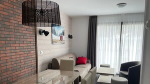 Una habitación de hotel moderna con pared de ladrillo, un sofá con almohada roja, una mesa de vidrio para comer y una pequeña mesa redonda.