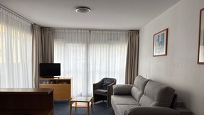 Una habitación de hotel moderna con sofá, televisión sobre un mueble de madera, una mesita y una silla.