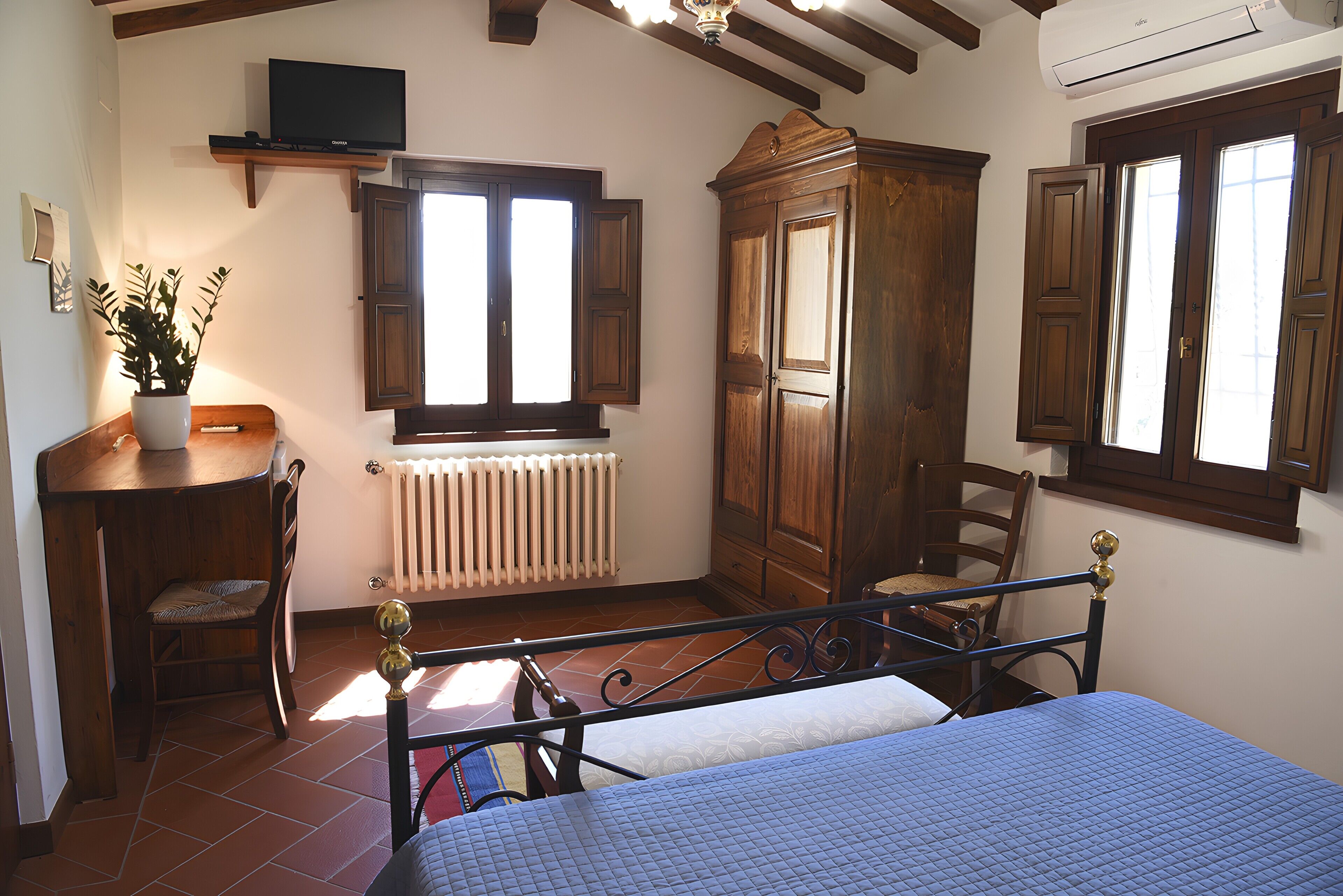 A bedroom with a bed, a wooden wardrobe, a radiator, a television on a shelf, and a window with shutters.