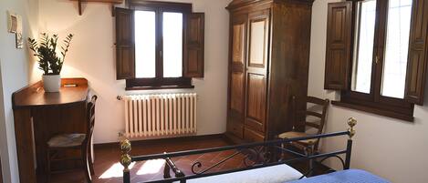 A bedroom with a bed, a wooden wardrobe, a radiator, a television on a shelf, and a window with shutters.