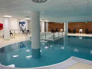 Piscina interna com água cristalina, rodeada por cadeiras de descanso e uma parede de madeira.