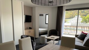 Et hotelværelse med spisebord, to stole, en sofa, et lille bord, et tv ophængt på væggen, et klædeskab og et vindue med udsigt til bygninger og træer.