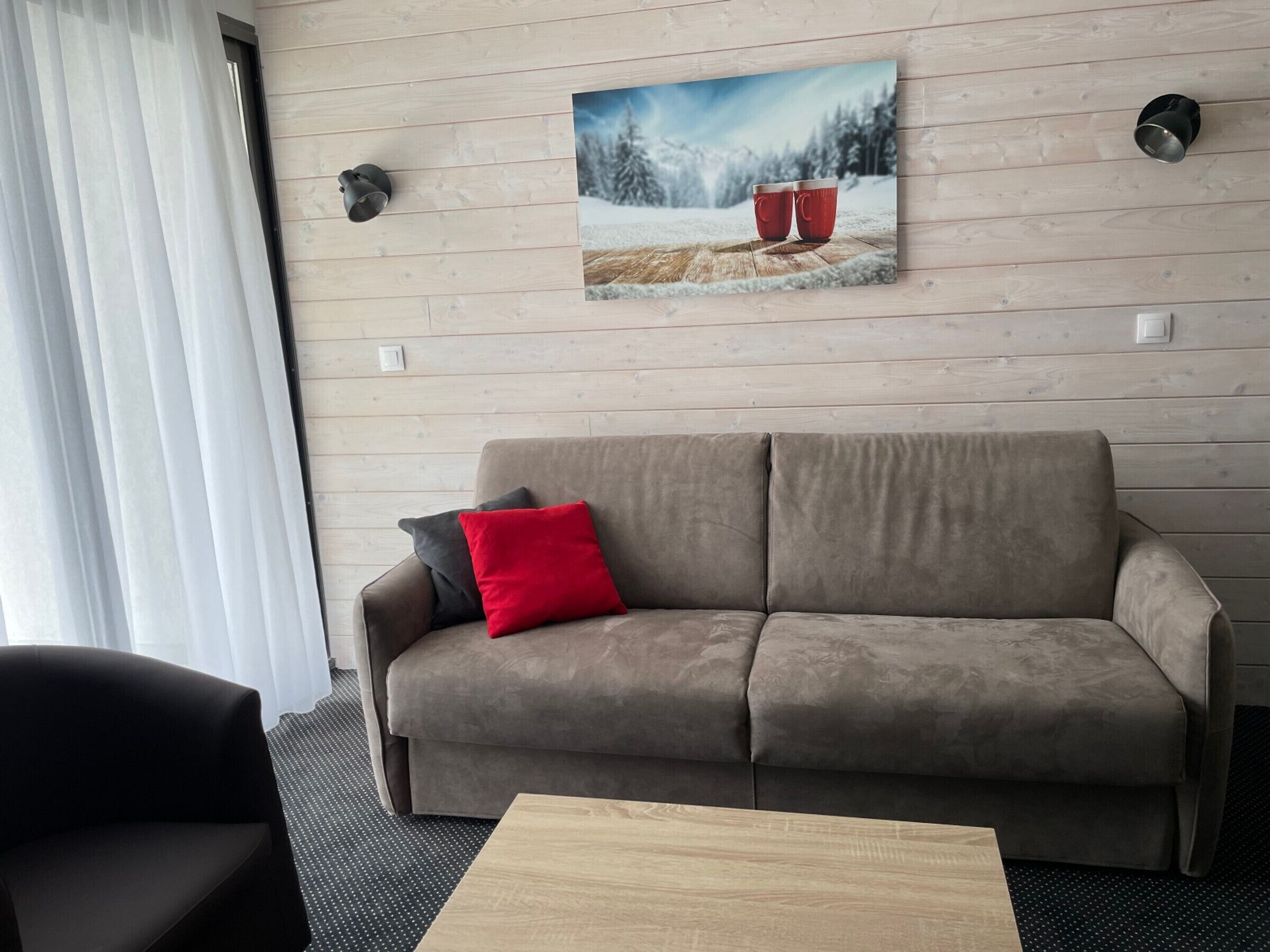 Un salon moderne avec un canapé gris, un coussin rouge et une table basse en bois. Un tableau représentant un paysage enneigé est accroché au mur.