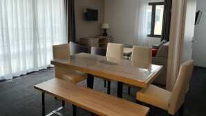 Ein Hotelzimmer mit Essbereich, ausgestattet mit einem Holztisch und -stühlen, einer Bank, einem an der Wand befestigten Fernseher und einem Fenster mit Vorhängen.