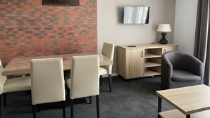 Hotellihuone, jossa on yksi seinä koristeltu tiilillä, taulu-tv, työpöytä lampun kanssa, ruokapöytä tuoleineen ja sohva.