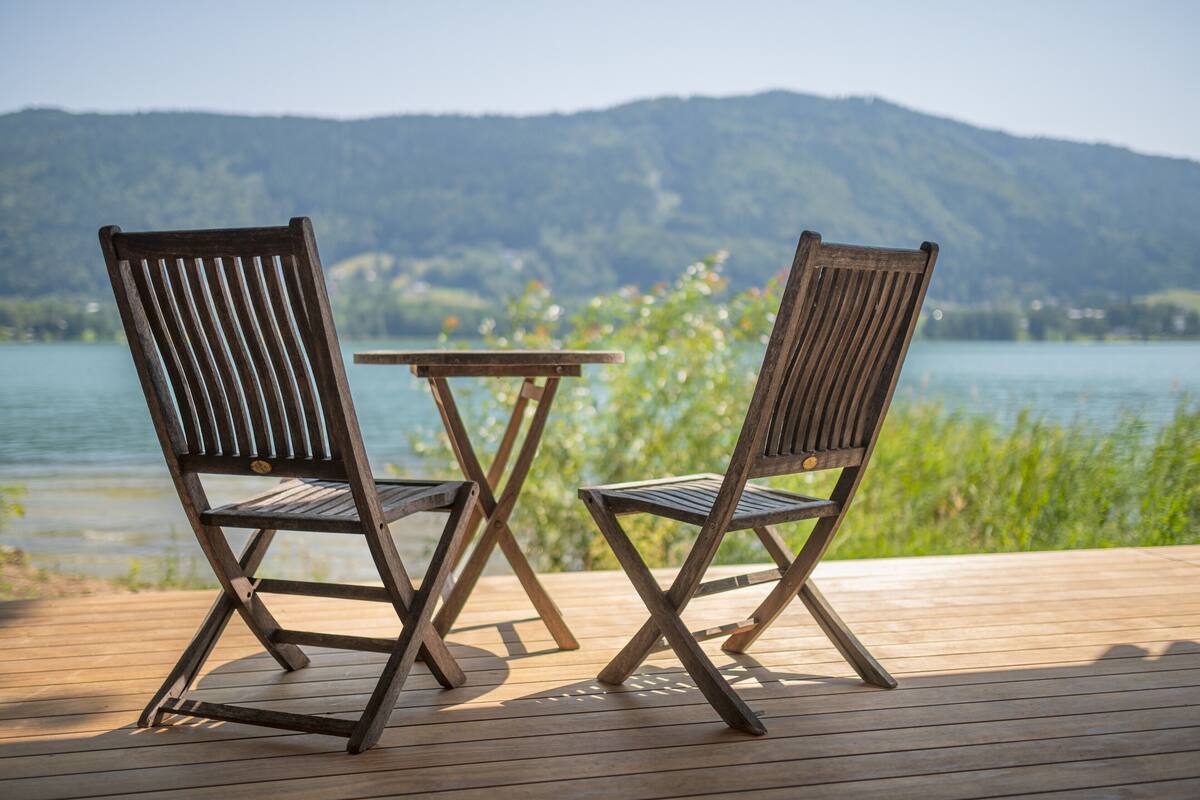 Two wooden chairs and a small table on a wooden deck with a lake and mountains in the background.