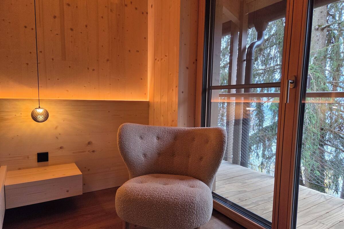 A cozy room with a plush armchair, a wooden bench, and a large window offering a view of the forest.