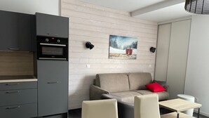 Cocina moderna con horno empotrado y un mueble gris. Hay un sofá color beige con un cojín rojo y una pequeña mesa con una silla. Un cuadro de un paisaje nevado adorna la pared.