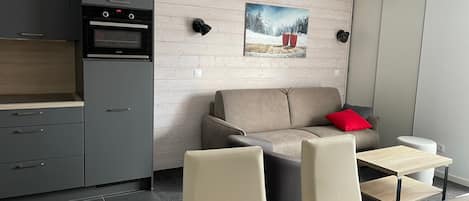 A modern kitchen with a built-in oven and a grey cabinet. There is a beige sofa with a red pillow and a small table with a chair. A painting of a snowy landscape is on the wall.
