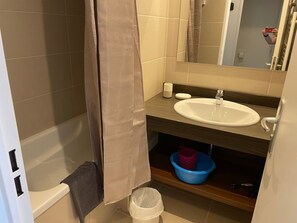 Un baño con bañera, lavamanos, espejo y repisa con una cesta azul y un cubo de basura blanco.