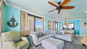 A living room with a sectional sofa, a patterned armchair, a ceiling fan, and a view through a large window. - Summer House 406B (Orange Beach)