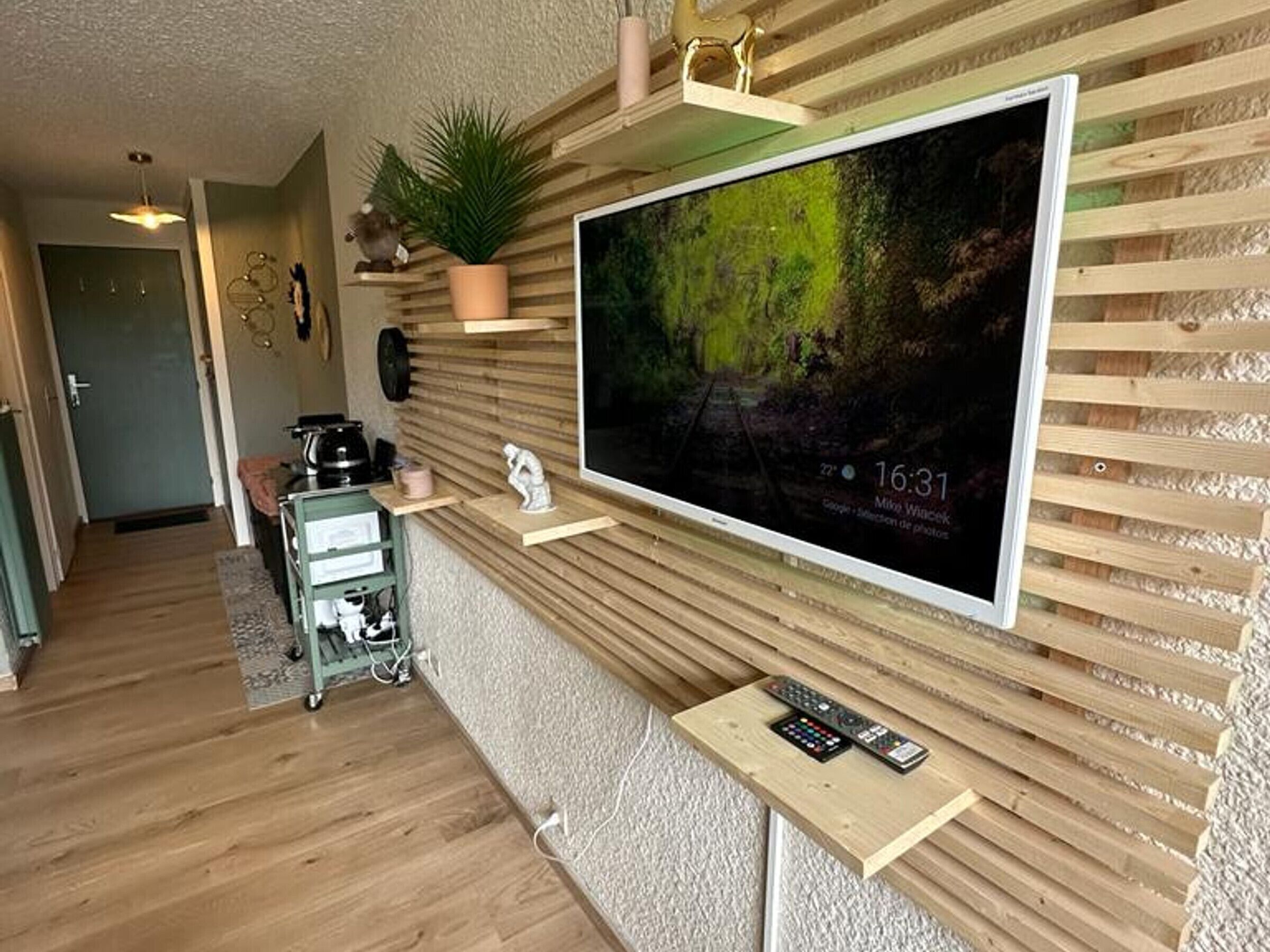 A living room with a large flat-screen TV mounted on a wooden wall, a small shelf with decorative items, and a remote control on a wooden stand.