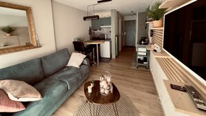 Een moderne woonkamer met een grijze bank, een salontafel met decoratieve objecten, een televisie en een keuken op de achtergrond.