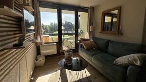 Een woonkamer met een groene bank, een klein bijzettafeltje en uitzicht op een tuin en bergen door grote ramen.