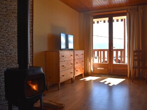 A cozy room with a wood-burning stove, a wooden dresser, a television, and a balcony with a view. - Charming South-Facing 2-Rooms with Garage and Balcony (Font-Romeu-Odeillo-Via)