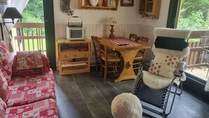 Een gezellige kamer met een houten tafel, een bank en een stoel. Er is een plank met decoratieve objecten en een raam met uitzicht op groen.