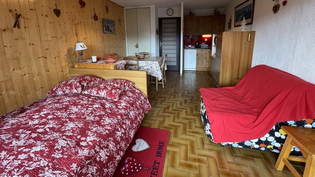 Une pièce chaleureuse aux murs en bois, avec un lit recouvert d’une courtepointe à motifs floraux rouges, un canapé rouge, une petite table en bois et, au fond, un coin cuisine.