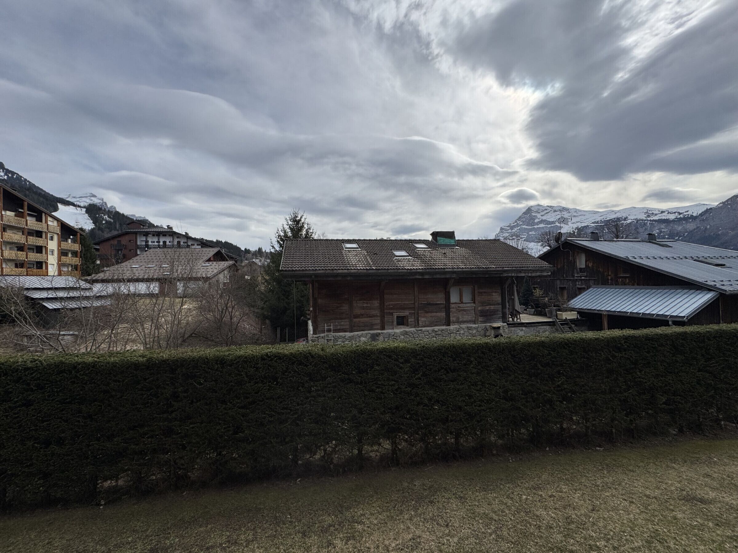 Un bâtiment traditionnel en bois, doté d’un toit en pente, entouré d’une haie et avec une chaîne de montagnes en arrière-plan.