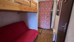 Une petite pièce étroite, avec un lit simple, un lit superposé en bois au-dessus, un rideau à motifs et une porte dotée d’une poignée.
