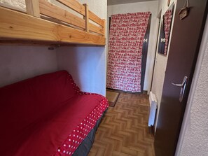 Ein kleiner, schmaler Raum mit einem Einzelbett und einem darüberliegenden Holzbett, einem gemusterten Vorhang und einer Tür mit Griff.