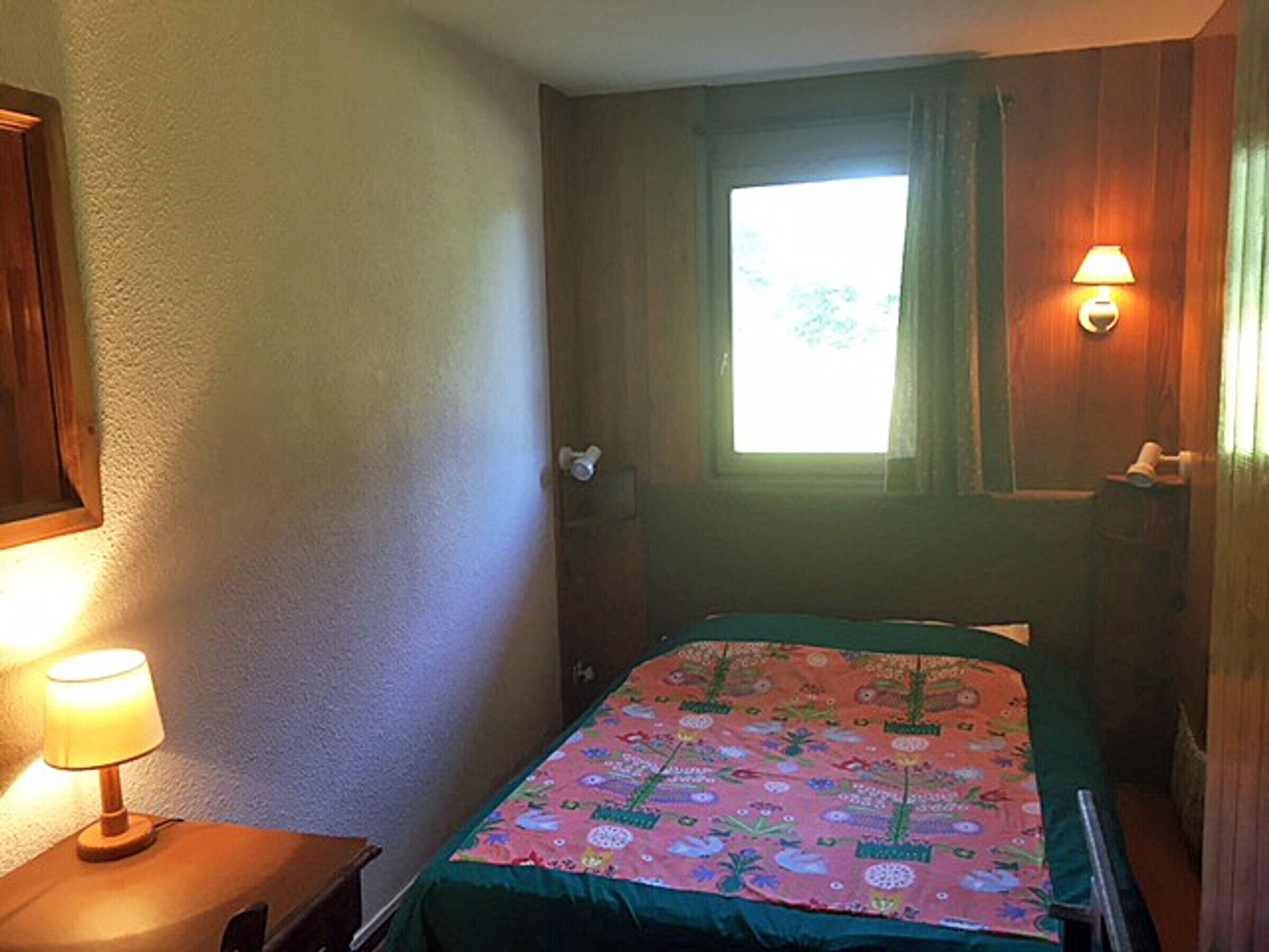 A small, dimly lit room with a single bed, a nightstand with a lamp, and a window with curtains.