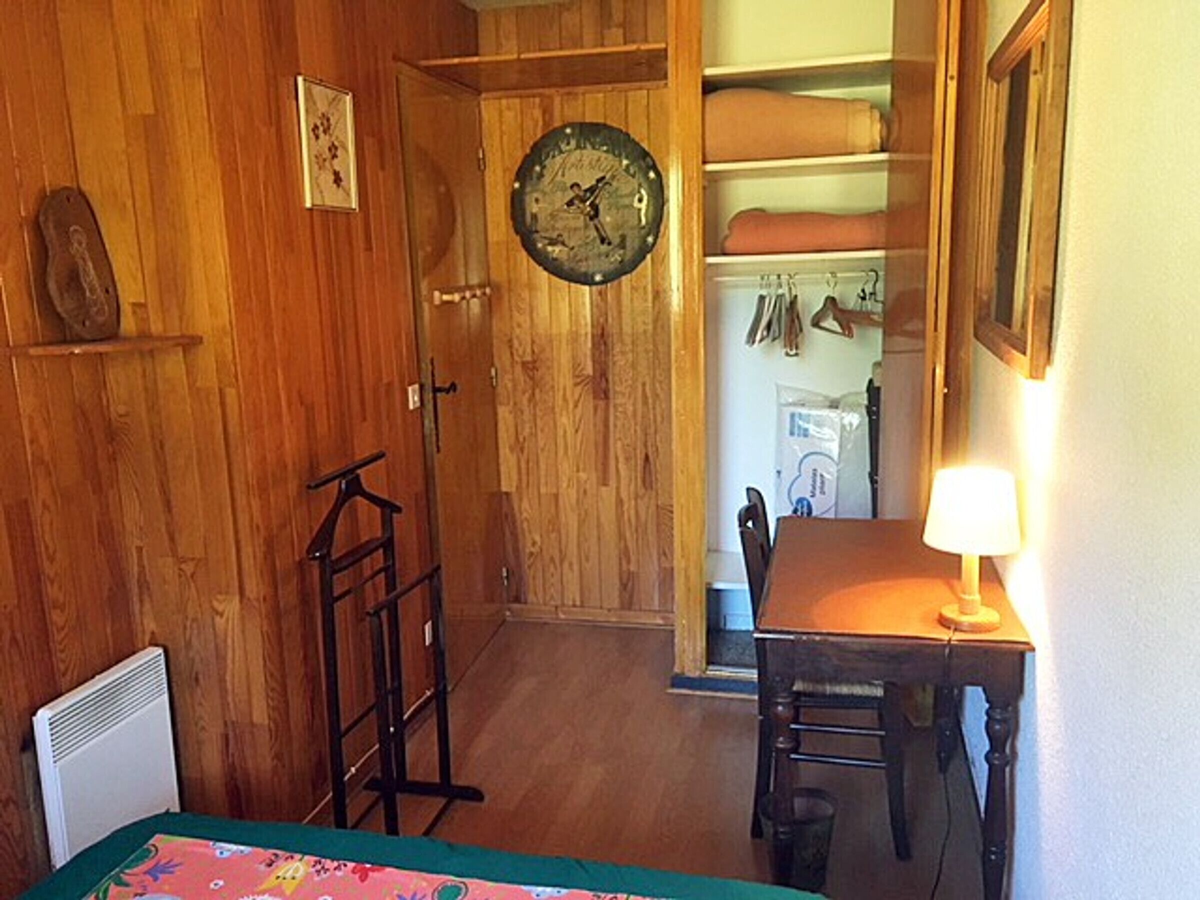 A wooden-paneled room with a bed, a table with a lamp, a chair, a coat rack, and a wardrobe with hanging clothes.