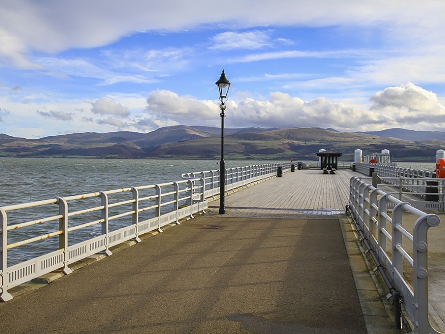 A pier with a metal railing, a lamp post, and a view of the water and distant hills.