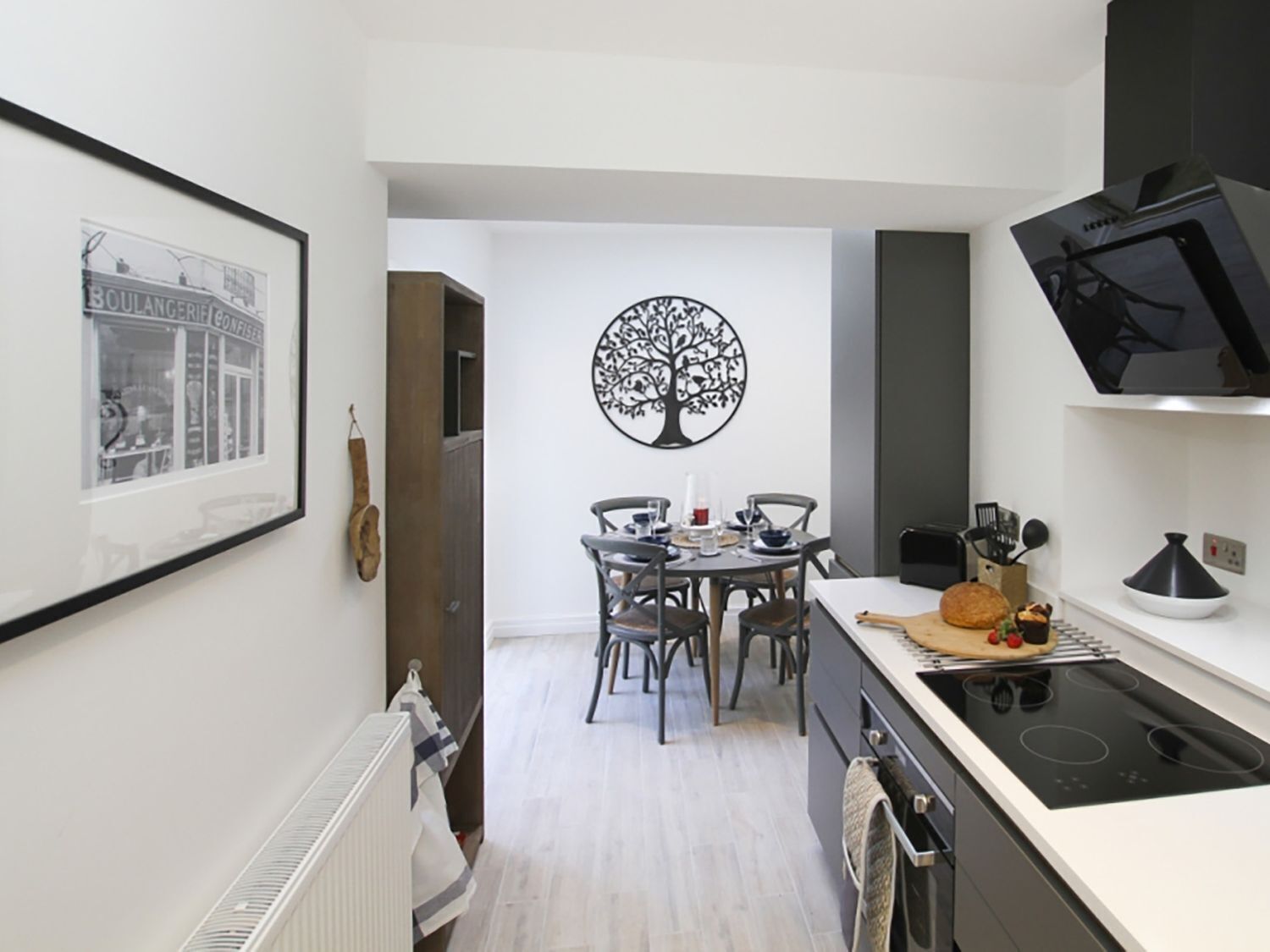 A modern kitchen with a dining area, featuring a black tree wall art, a framed picture, and a black TV on the wall.