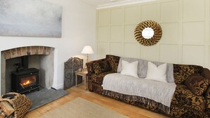 A cozy living room with a fireplace, a patterned sofa, a small table with a lamp, and a decorative wall hanging. - Tri Raglan Bach (Beaumaris)