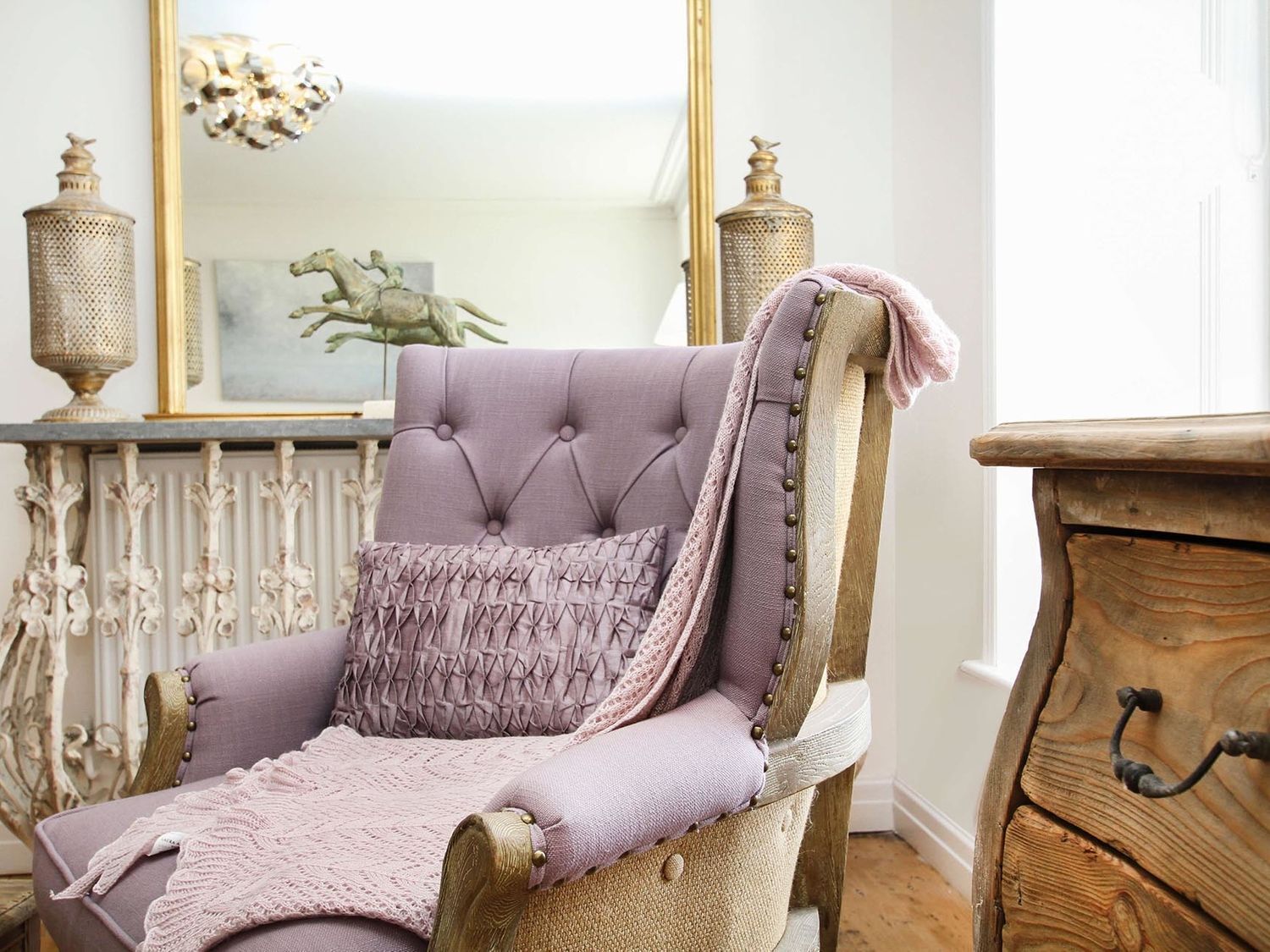 Een gestoffeerde fauteuil met een gebreide plaid en een kussen, geplaatst naast een houten dressoir en een spiegel met decoratieve objecten.