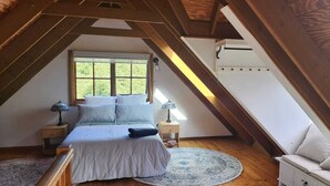 A bedroom with a sloped ceiling, a bed with pillows, a nightstand with a lamp, and a window with a view of greenery. - Cottage in beautiful Blue Mountains (Mount Victoria)