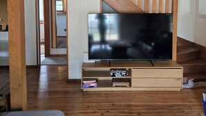 A spacious living room with a large flat-screen TV on a wooden stand, a staircase with wooden railings, and a cozy seating area with a grey sofa and a green rug. - Cottage in beautiful Blue Mountains (Mount Victoria)