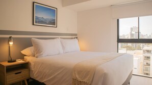 A hotel room with a bed, a nightstand, a lamp, a framed picture on the wall, and a window with a city view. - w Enchanting 1BR w Balcony in Barranco (Lima)