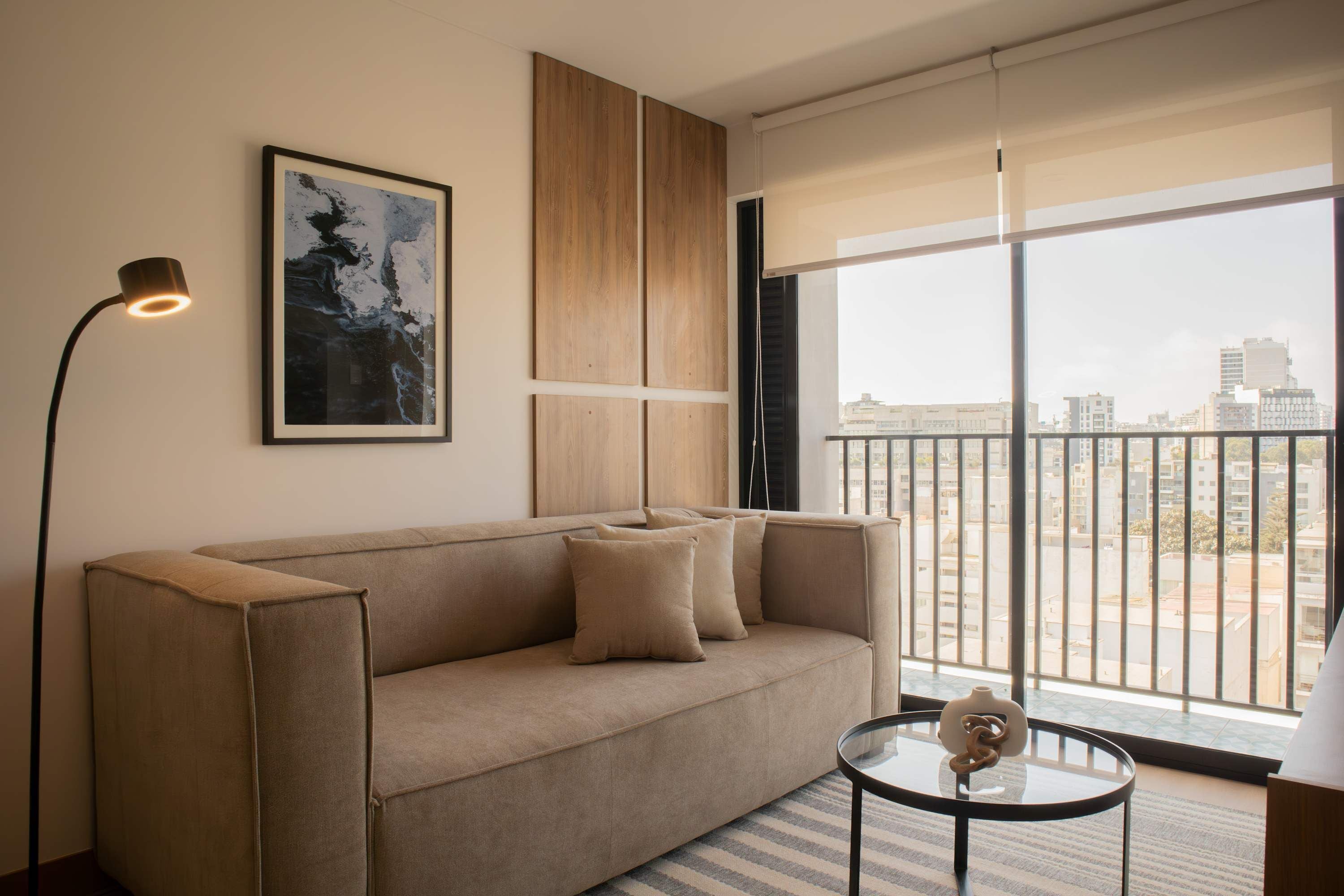A modern living room with a beige sofa, a glass coffee table, a floor lamp, and a large window offering a city view.