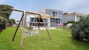 A house with a swing set in the yard, a white fence, and a blue building in the background. - Holiday Home “Les Sables Pailletés 14p” with Sea View, Shared Terrace & Wi-Fi (Les Sables-d'Olonne)