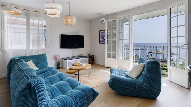 Un salón espacioso con un televisor grande, dos sofás azules, una mesita y un balcón con vistas al mar.