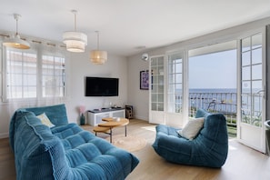 A spacious living room with a large TV, two blue sofas, a small table, and a balcony with a view of the sea. - Apartment 'Les Sables Pailletés 4p' with Sea View, Shared Garden and Wi-Fi (Les Sables-d'Olonne)