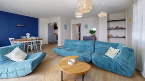 Una sala de estar con un sofá azul, una mesa de centro de madera y un comedor con una mesa y sillas blancas.