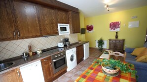 A compact kitchen with wooden cabinets, a microwave, a washing machine, and a dining area with a colorful tablecloth. - Rural Apartments La Viña (Apartment number 6) (Cangas de Onis)